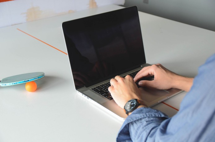 A person typing on a laptop in an office, with a ping pong paddle and ball nearby.