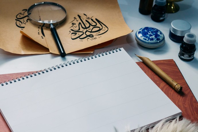 Creative calligraphy setup with Arabic script papers, magnifying glass, ink bottles, and blank notepad.